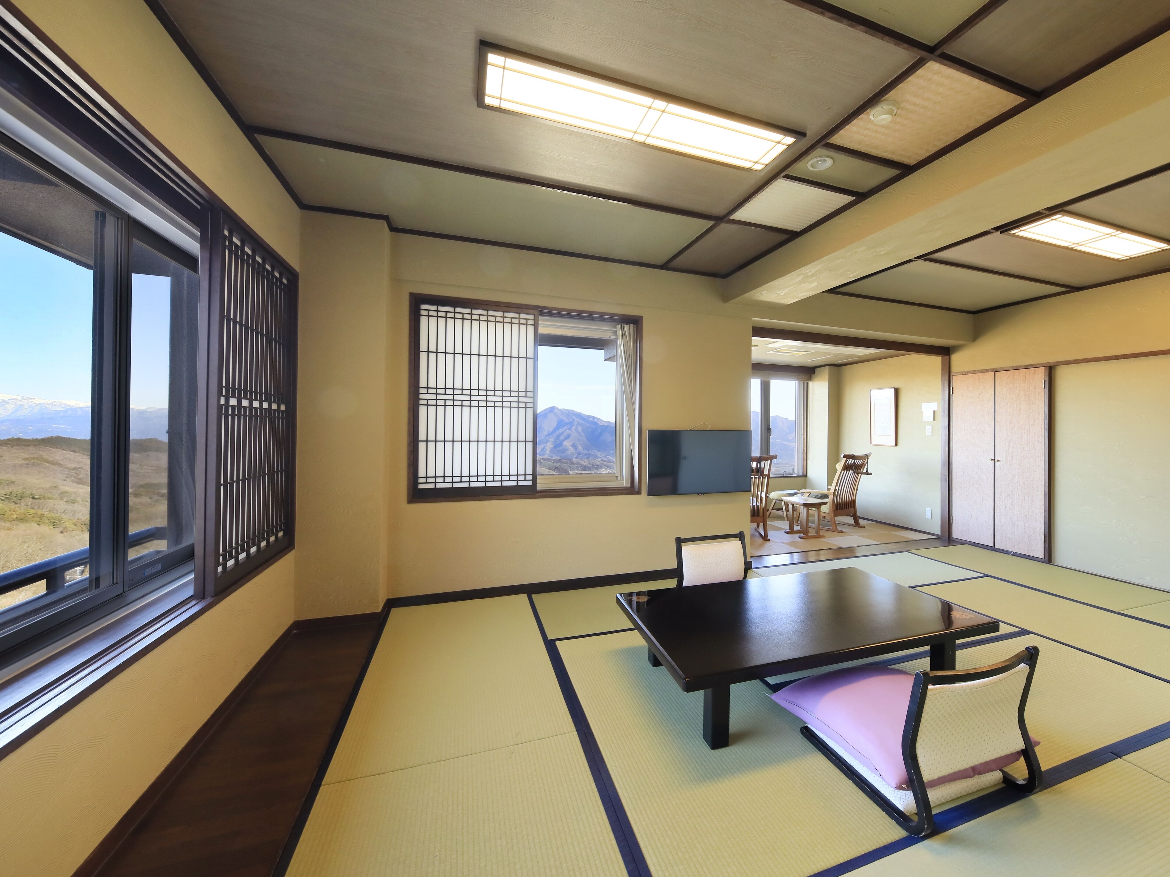 A tatami mat room with a tatami mat table, a television mounted on the wall, and a view of mountains through the window.