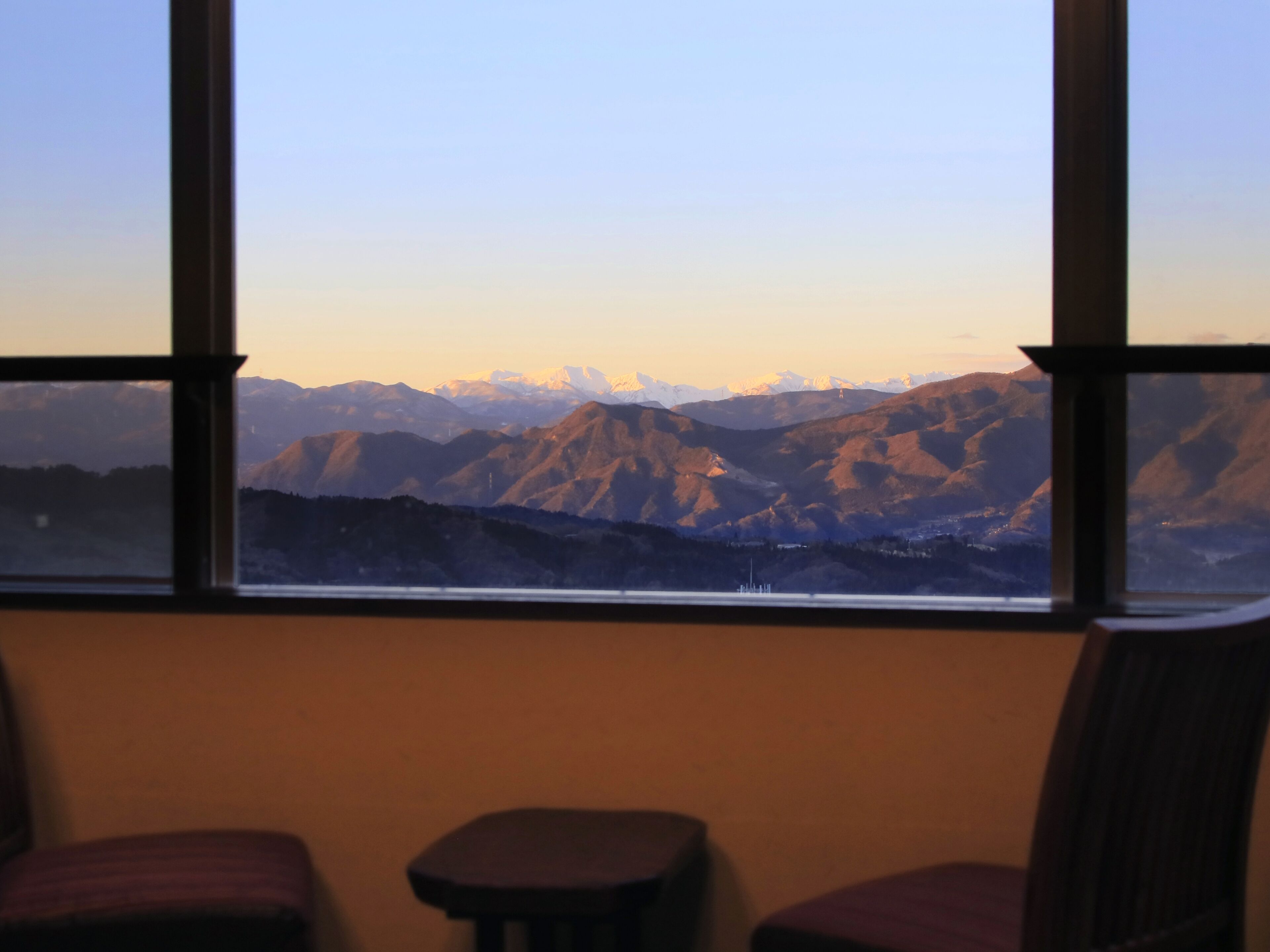 A room with a large window offering a view of mountains. There are two chairs and a small table in the foreground.