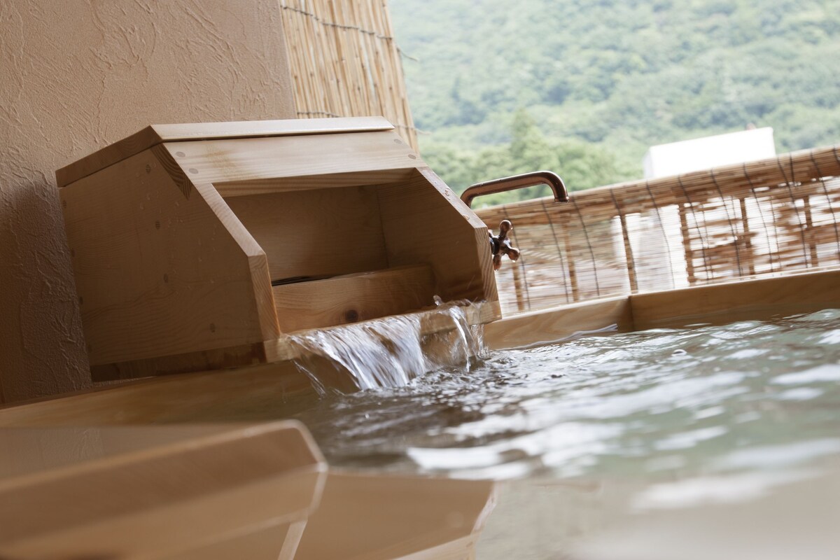 Un baño de aguas termales de madera con una cascada, rodeado de paredes de bambú y vistas a una montaña boscosa.
