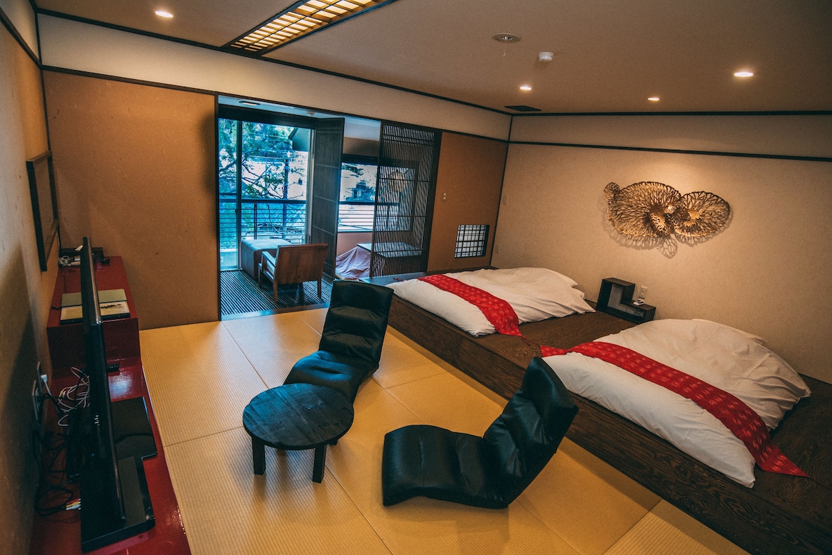 A hotel room with a large sliding door leading to a balcony, two beds with red and white bedding, a black chair, and a small table with a chair.