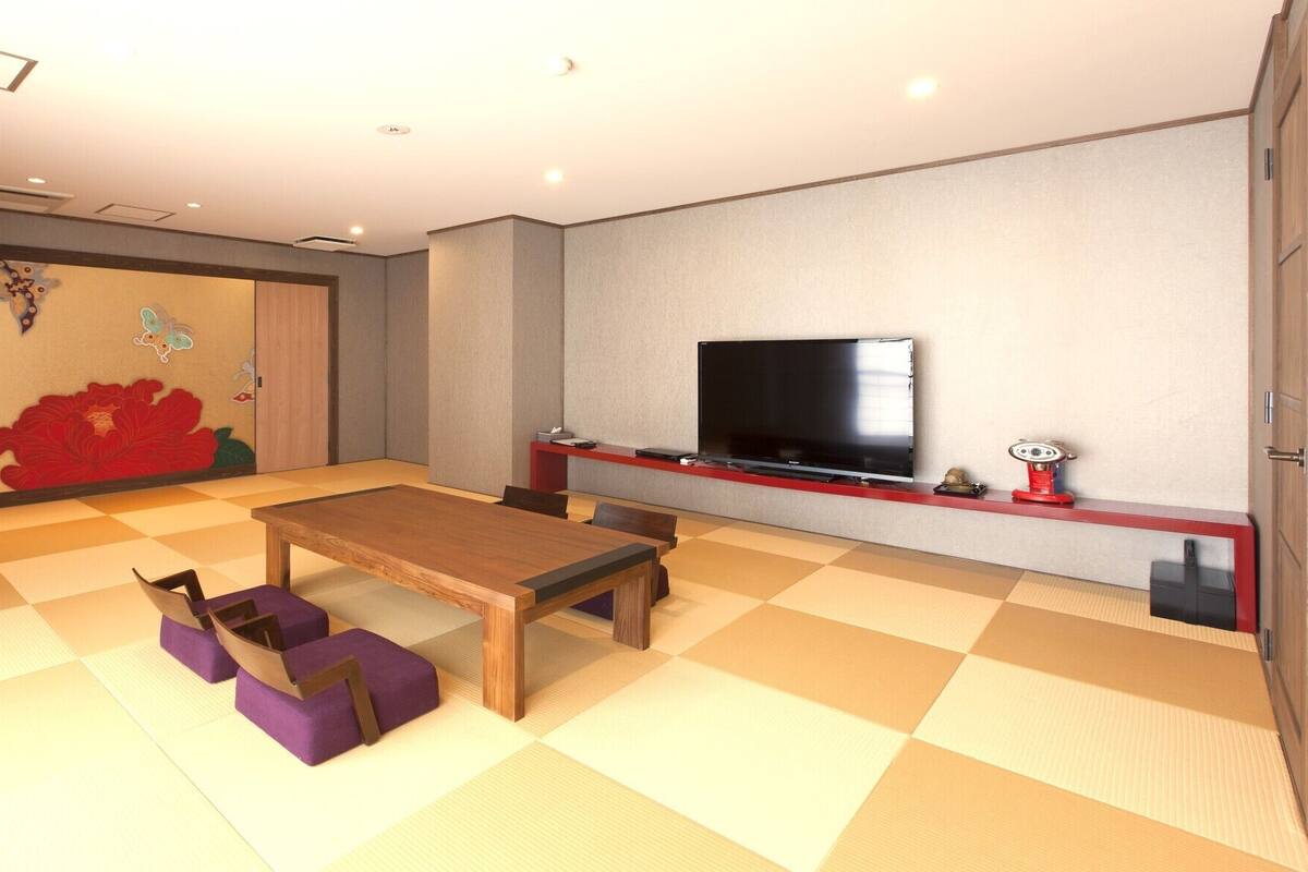 Una habitación con suelo de tatami, una mesa de madera, cojines morados, un televisor de pantalla plana y un mueble de entretenimiento rojo.