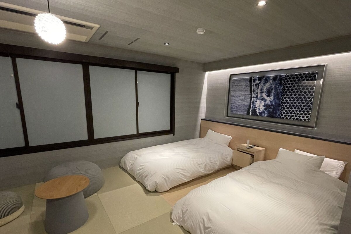 A modern hotel room with two beds, a small round table, and a large window with a view of a building.