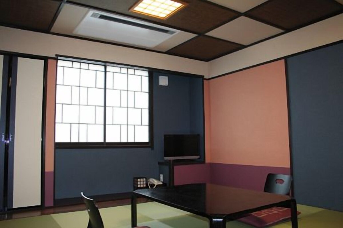 A room with a tatami mat floor, a low table, a chair, a television on a stand, and a window with a grid pattern.