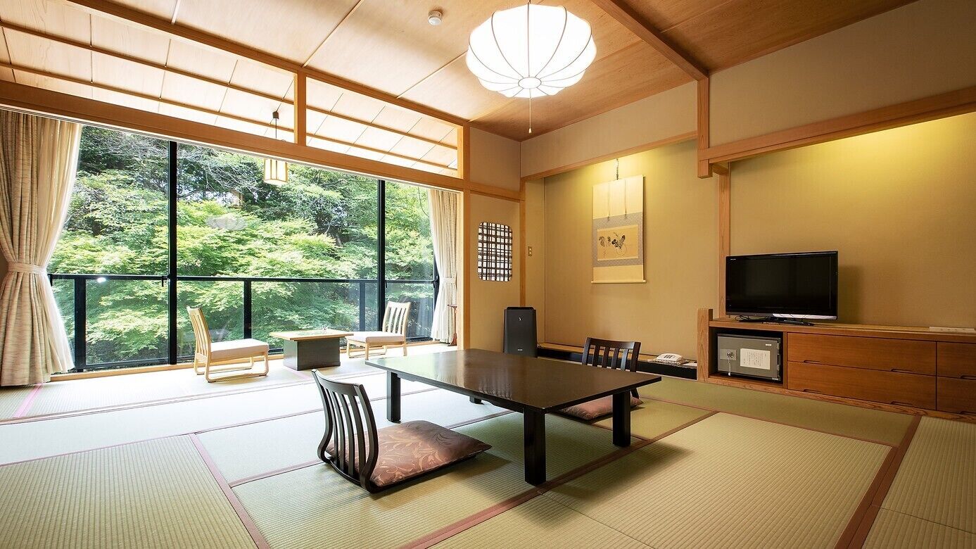 Une pièce de style japonais traditionnel, avec un sol recouvert de tatamis, une table basse, des chaises, un téléviseur et une vue sur la verdure à l’extérieur.
