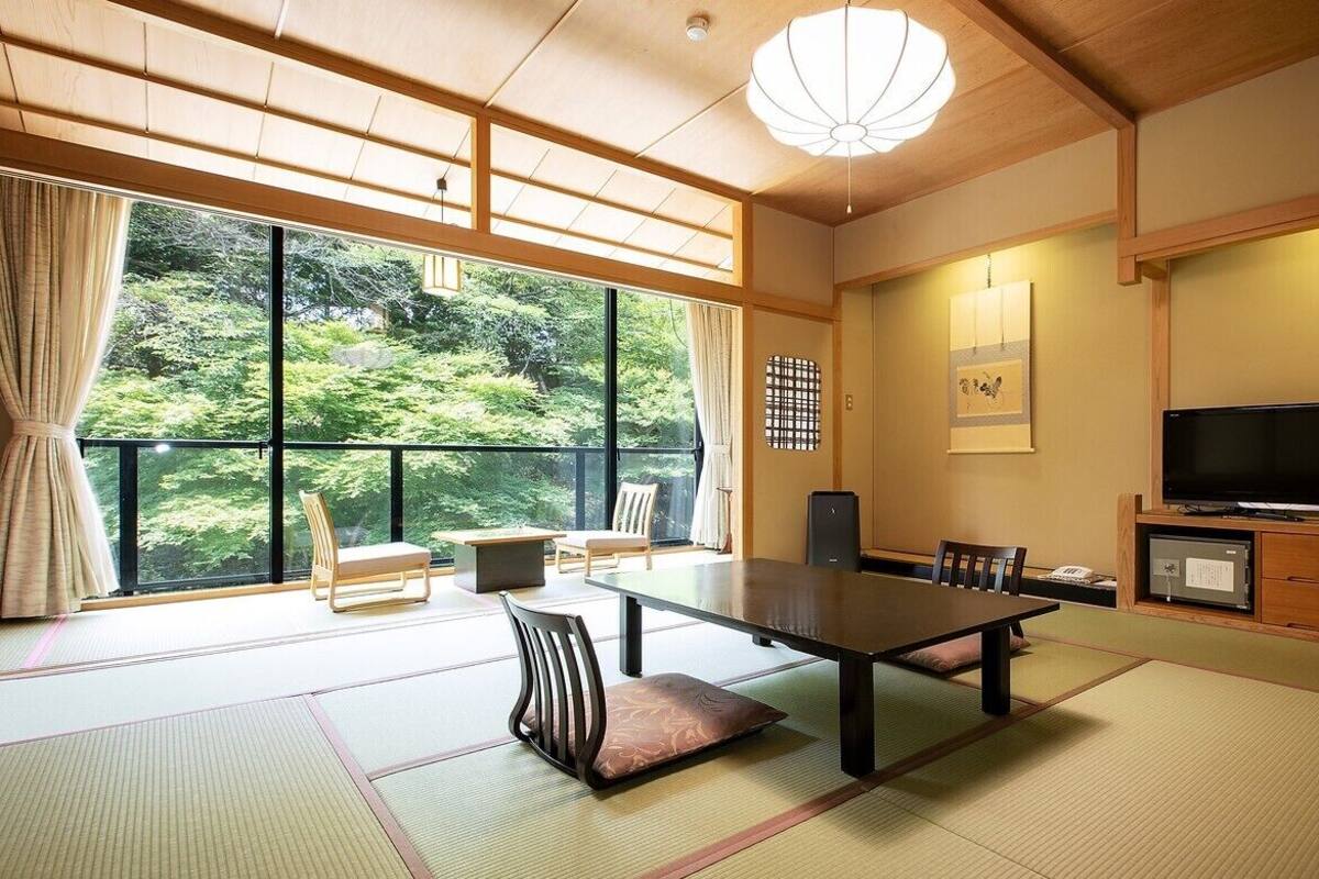 Une pièce de style japonais traditionnel, avec un sol recouvert de tatamis, une table basse, des chaises, un téléviseur et une vue sur la verdure à l’extérieur.