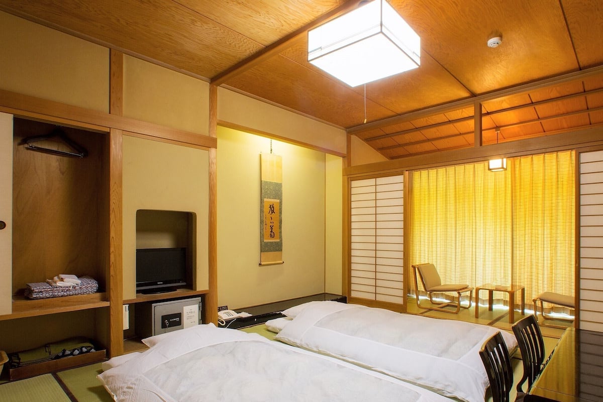 Une chambre d’hôtel avec deux lits, une télévision, une étagère avec des vêtements pliés, un bureau avec un téléphone et une porte coulissante donnant sur un balcon.
