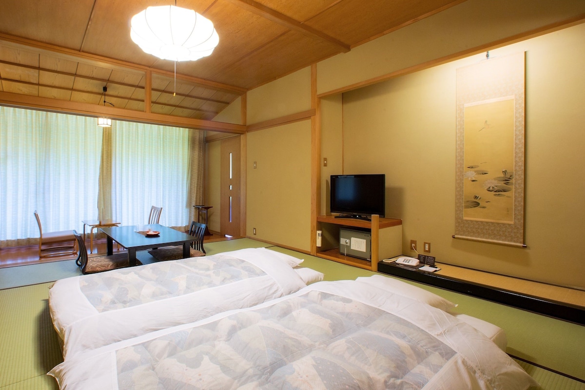 Quarto tradicional japonês com tatames, futon, televisão e mesa de jantar com cadeiras.