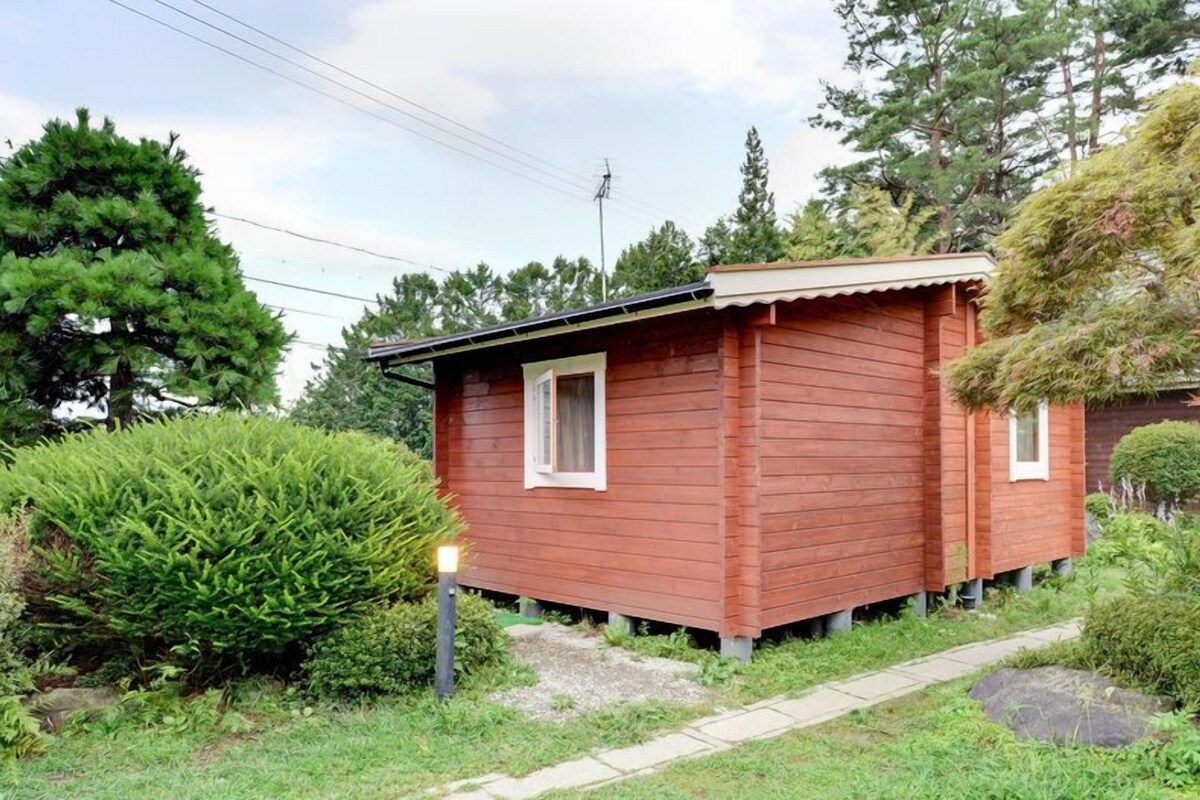 A small wooden cabin with a single window and a door, surrounded by lush greenery and trees.