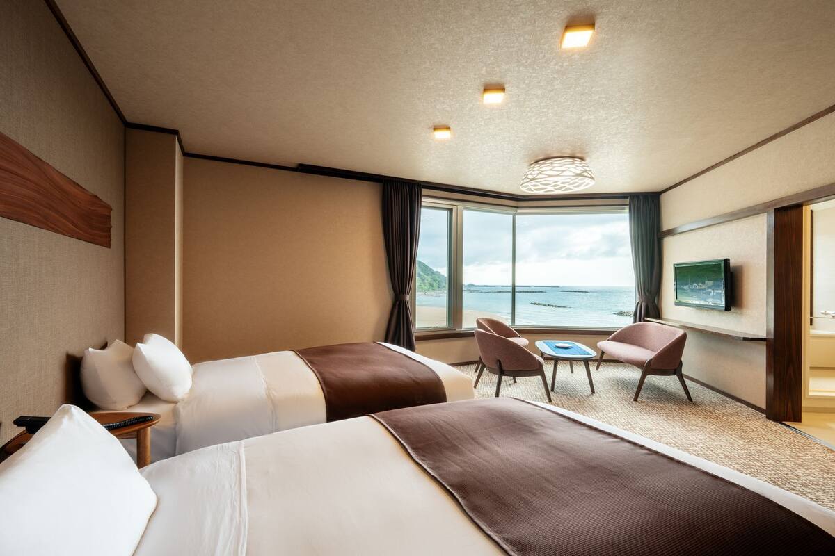 A hotel room with two beds, a TV, a desk with chairs, a large window with a view of the sea, and a shower area.