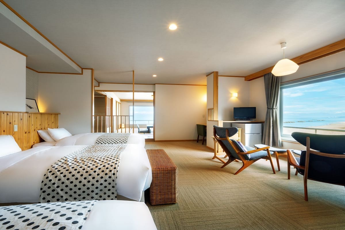 A hotel room with a bed, a desk with a TV, a chair, and a view of the beach.