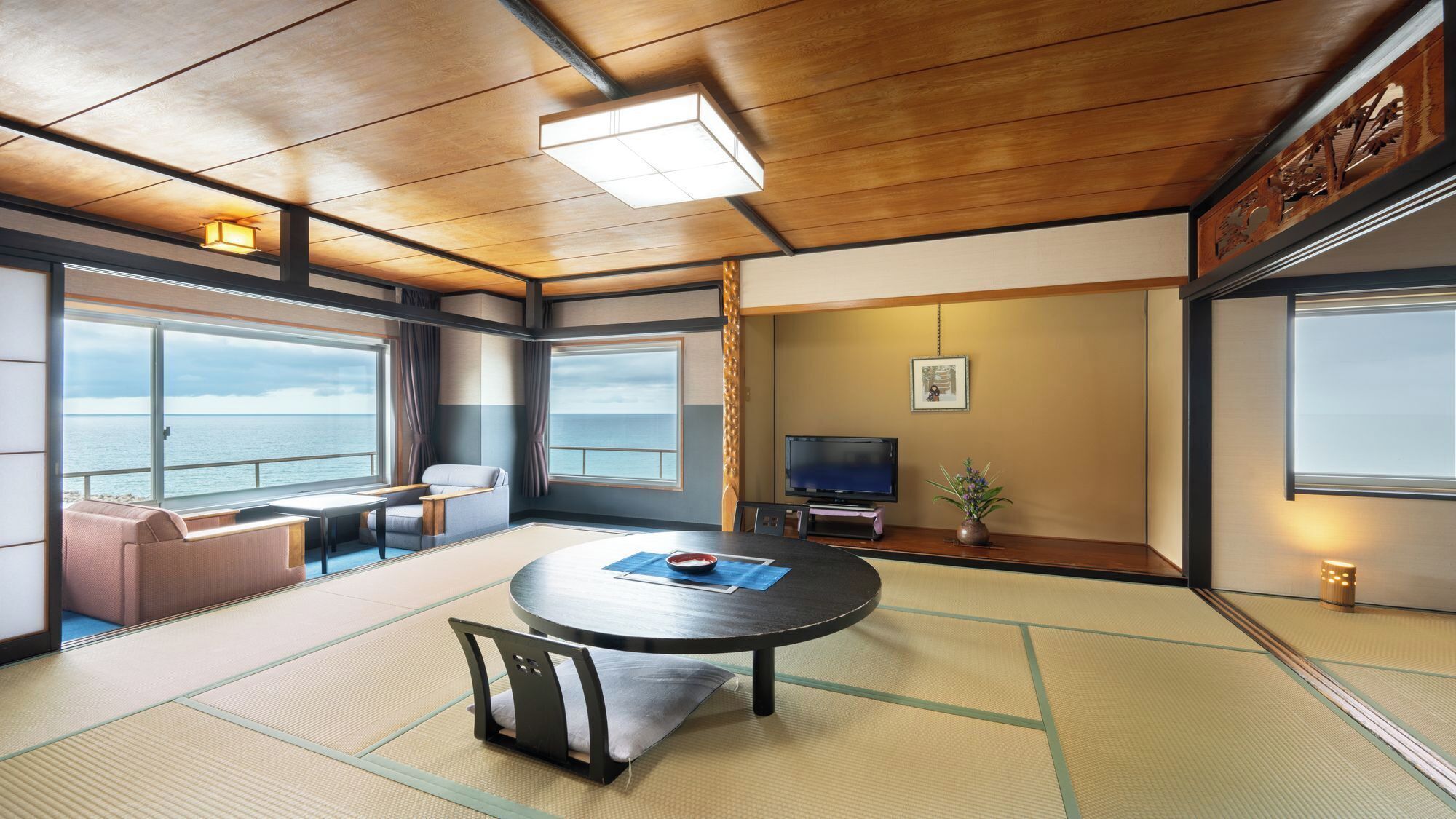 Una habitación con suelo de tatami, una mesa redonda, un sofá, un televisor y ventanas amplias con vista al mar.