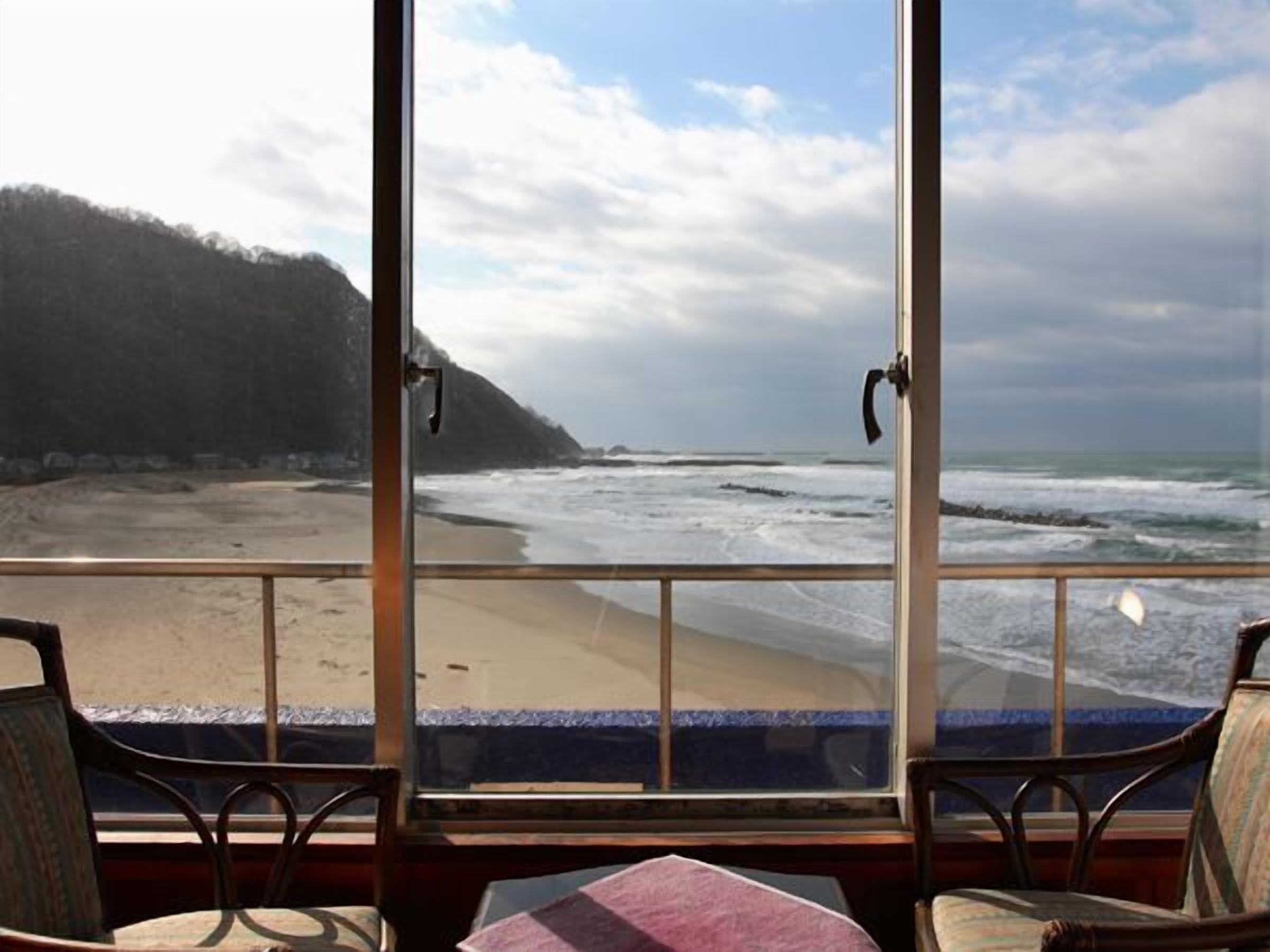 Una habitación con una amplia ventana que da a una playa y al océano, sillas de madera y una mesa.