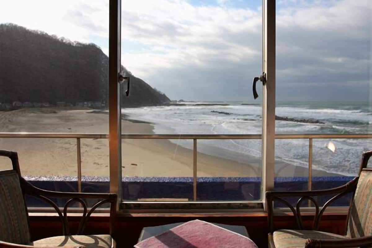 A room with a large window overlooking a beach and ocean, wooden chairs, and a table.