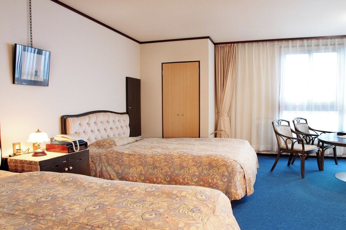 A hotel room with two beds, a desk, chairs, a lamp, and a wardrobe.
