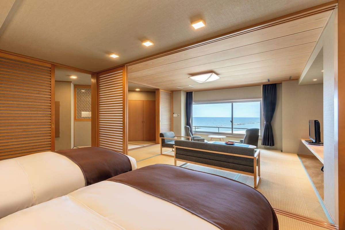 A hotel room with two beds, a desk with a chair, a small table, and a view of the sea.