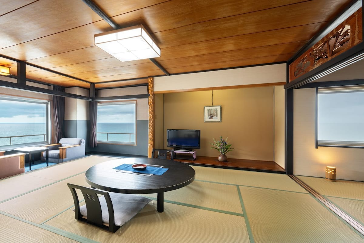 A room with tatami flooring, a round table, a sofa, a TV, and large windows with a view of the sea.