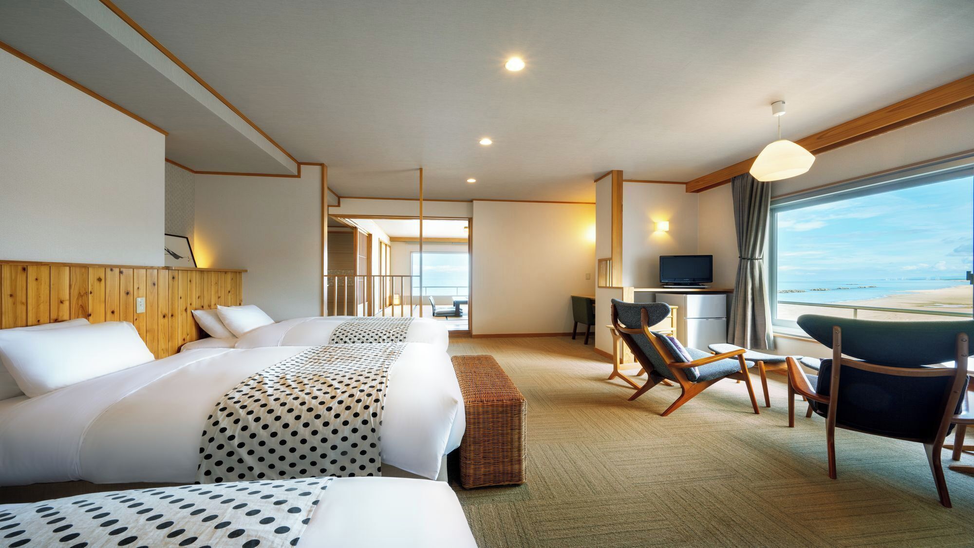 Habitación de hotel con una cama grande, un escritorio con televisor, una silla y vista a la playa.