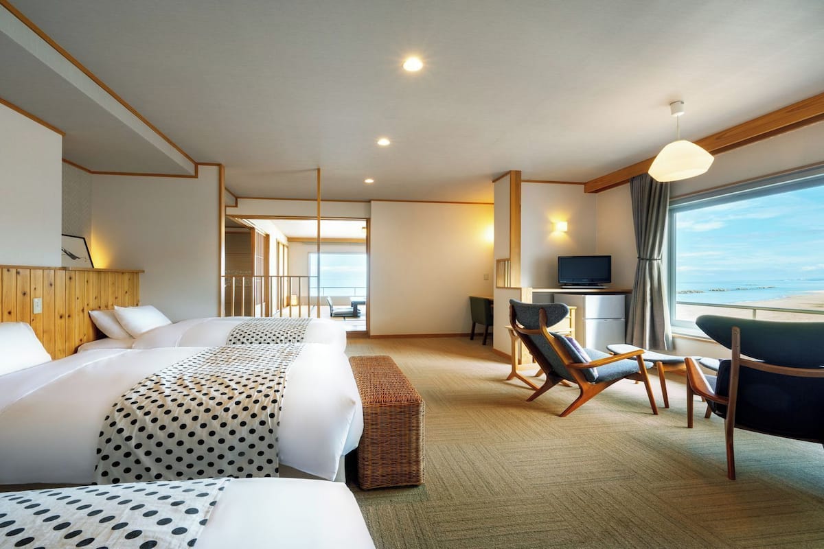 A hotel room with a large bed, a desk with a TV, a chair, and a view of the beach.