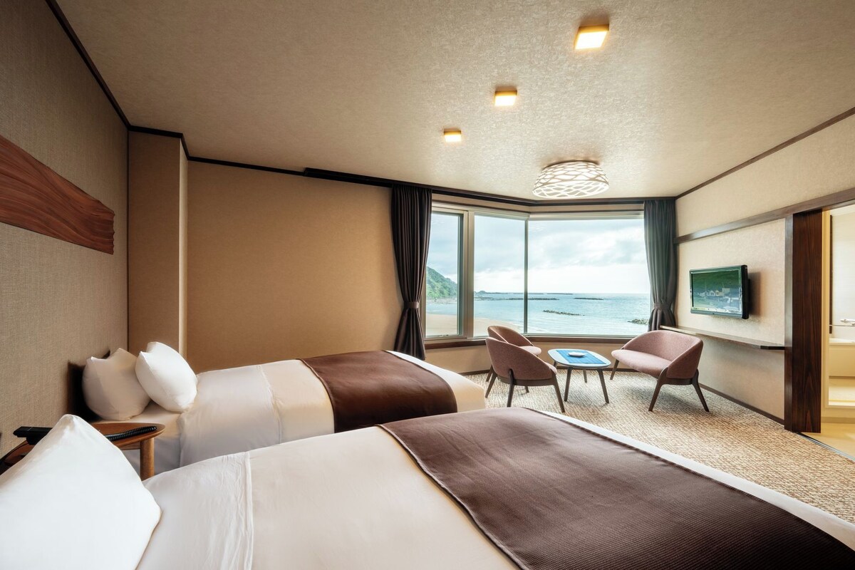 A hotel room with two beds, a small table with chairs, a TV, and a large window offering a sea view.