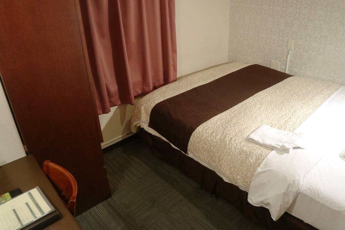 Une chambre d’hôtel avec un lit simple, un bureau avec une chaise et une fenêtre avec des rideaux.