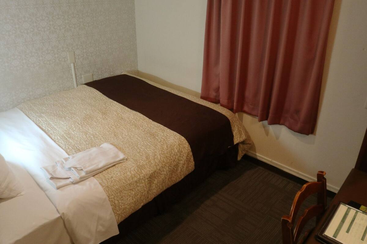 Un petit lit bien fait, avec une couverture marron et beige, un oreiller blanc et une serviette blanche pliée sur le côté gauche. À droite, on aperçoit une chaise en bois et une petite table sur laquelle est posé un menu ou un dépliant.