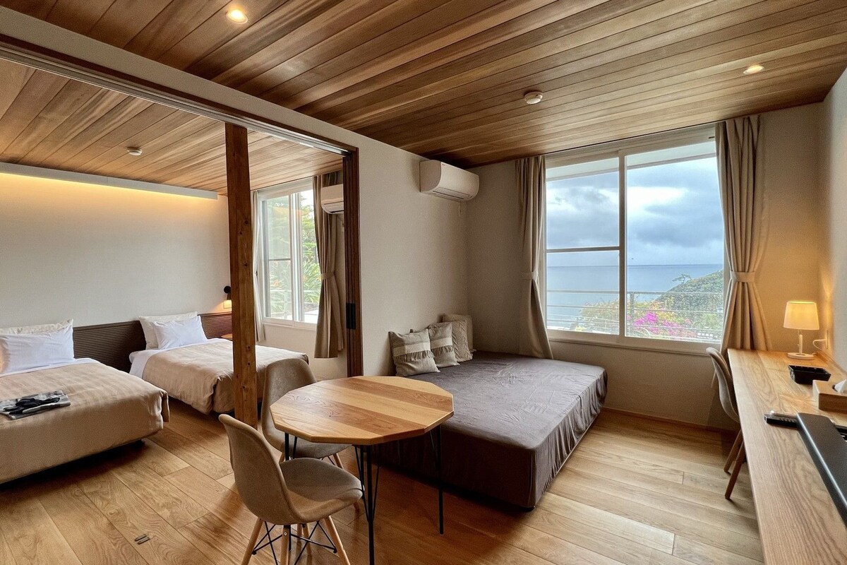 Una habitación de hotel moderna con techo de madera, un ventanal con vistas, un escritorio con lámpara y dos camas.