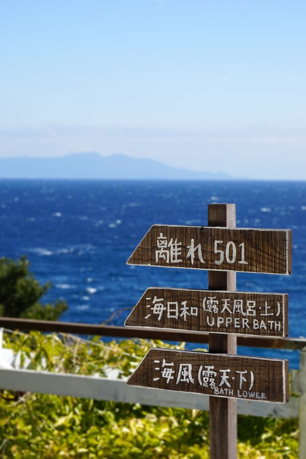 A wooden signpost with directions to Upper Bath and Bath Lower, set against a backdrop of the ocean and mountains. - Garden Villa Shirahama (Shimoda)