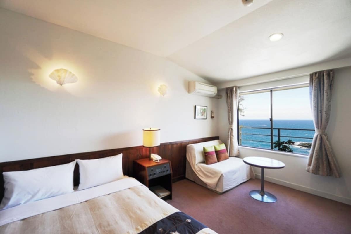A hotel room with a bed, bedside table, lamp, and a window with a view of the ocean.