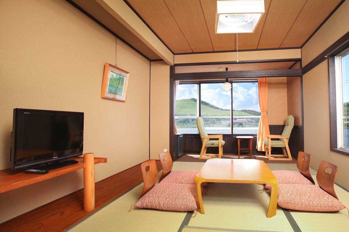 A room with tatami mats, a television, a table, chairs, and large windows with a view of hills.