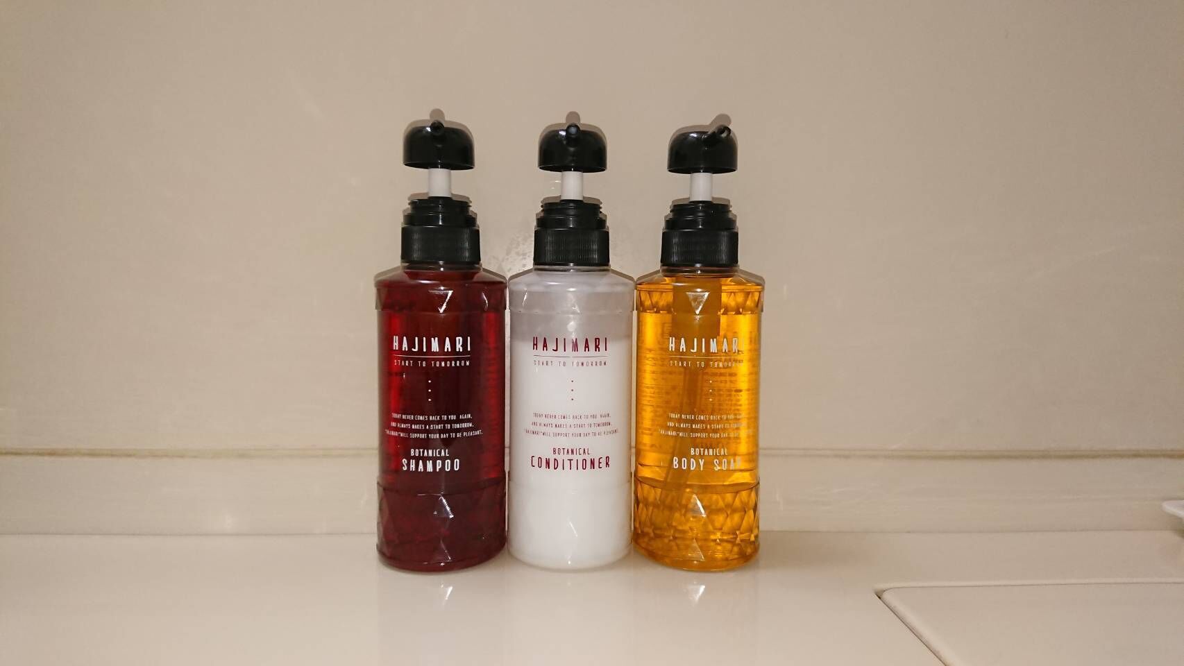 Trois produits de la gamme Hajimari : un flacon rouge contenant un shampooing à base de plantes, un flacon blanc contenant un après-shampooing à base de plantes, et un flacon jaune contenant un savon pour le corps à base de plantes.