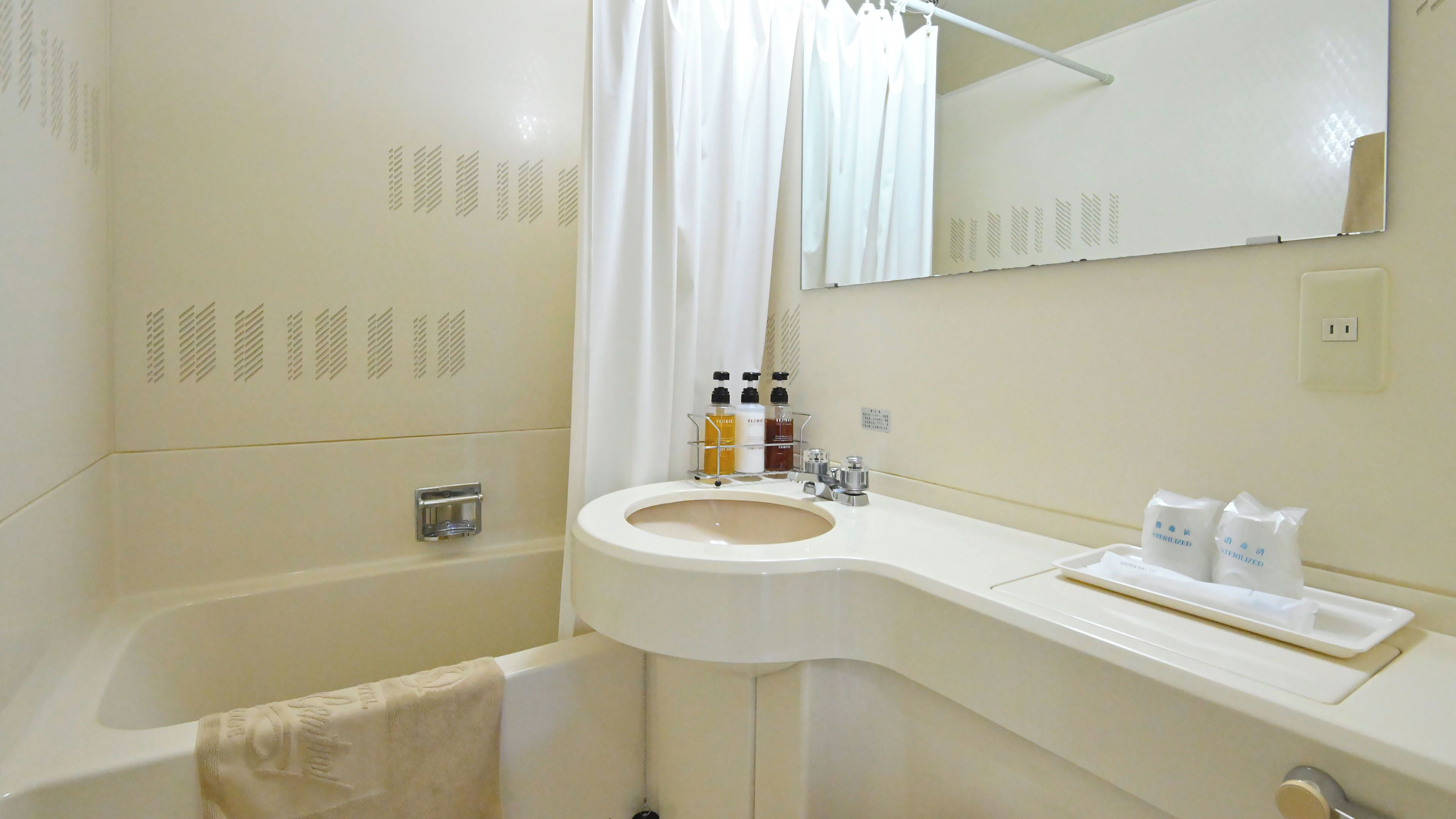 Une salle de bain avec un lavabo blanc, un miroir et une baignoire. On trouve des articles de toilette sur le lavabo et une serviette est accrochée au mur.