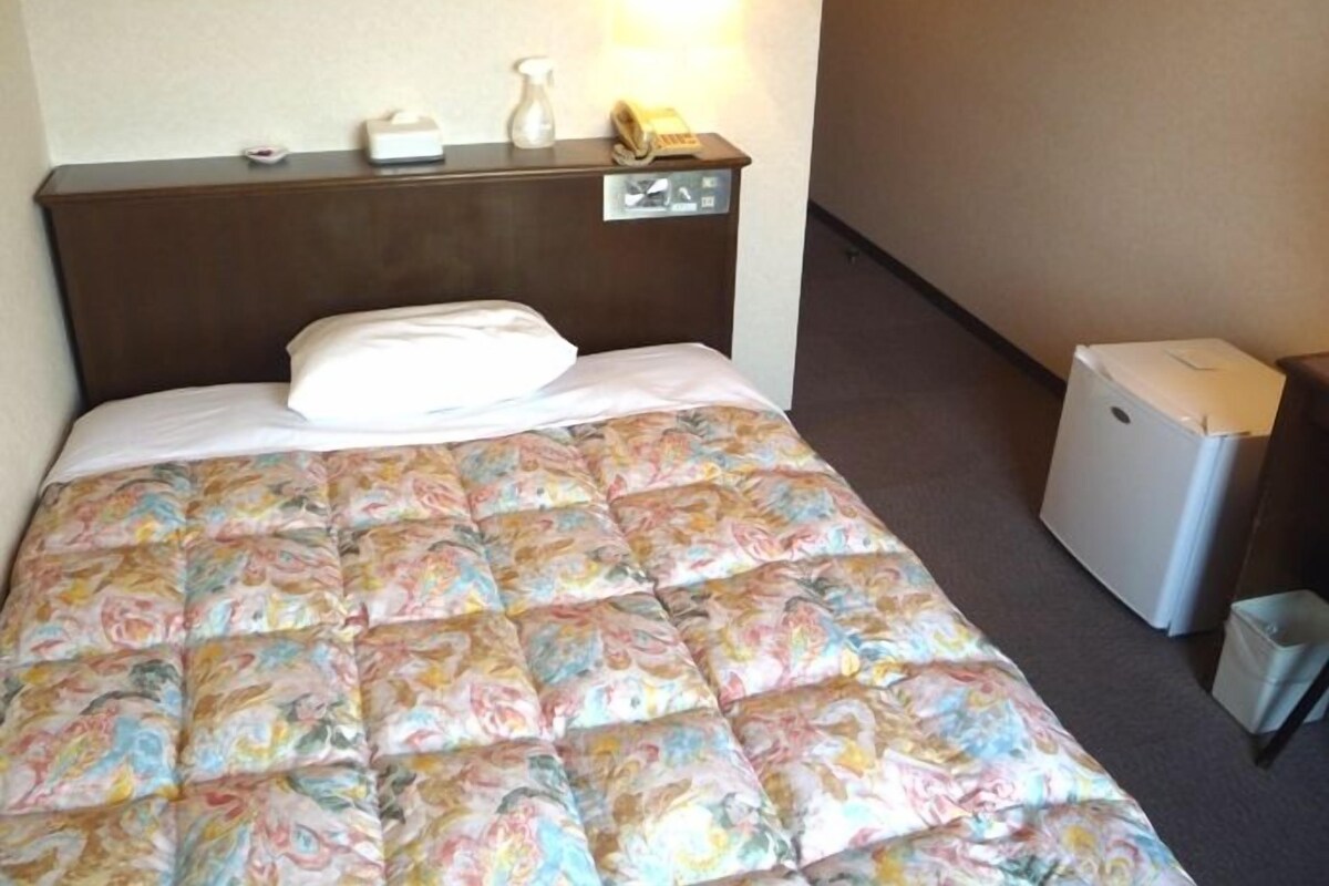 Una habitación de hotel con una cama, una mesita de noche, una lámpara, un cubo de basura y un refrigerador pequeño.