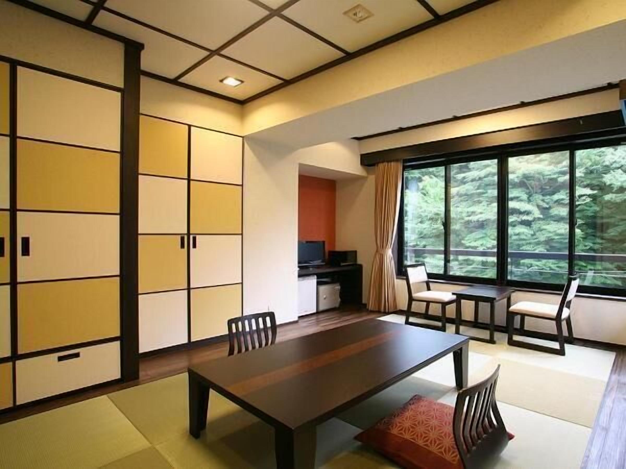 A room with a wooden dining table, chairs, a TV, and a large window with a view of greenery.
