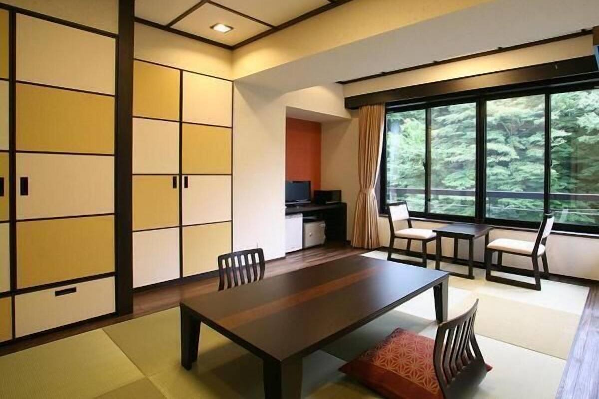 A room with a wooden dining table, chairs, a TV, and a large window with a view of greenery.