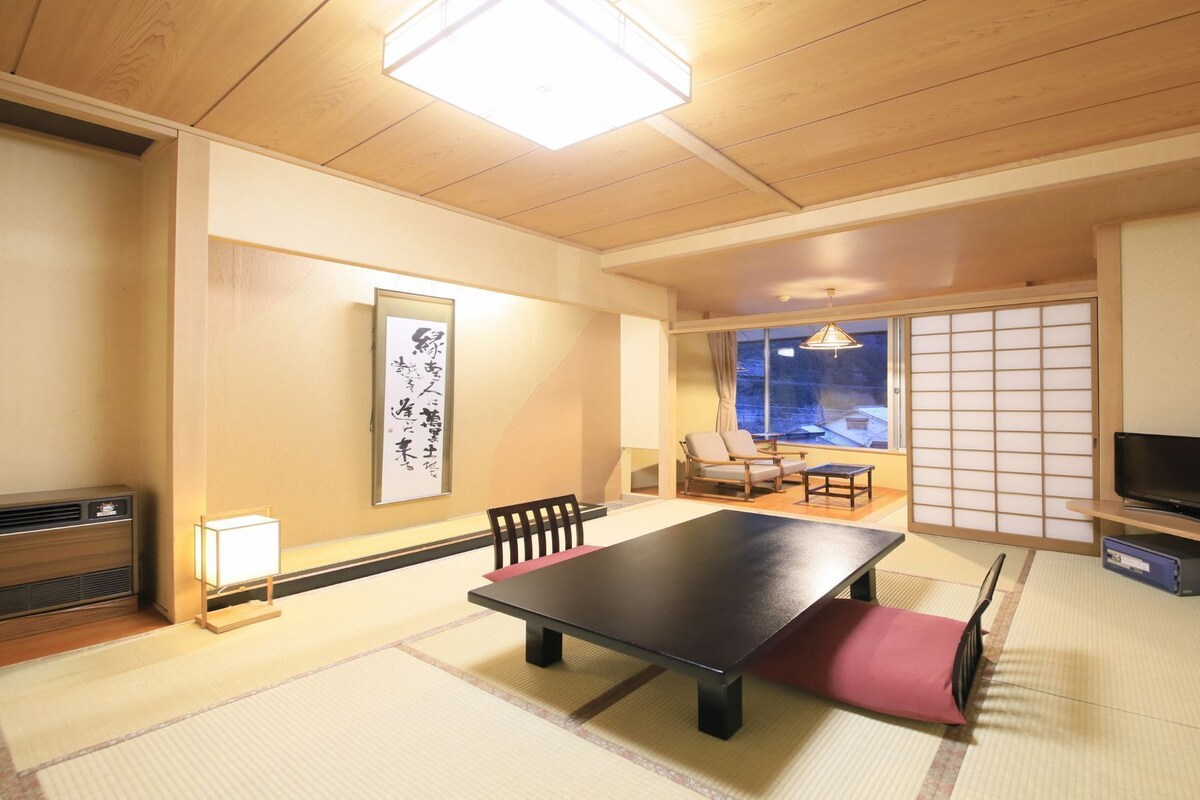 Una habitación japonesa tradicional con suelo de tatami, una mesa baja, sillas, un televisor y una puerta corredera.