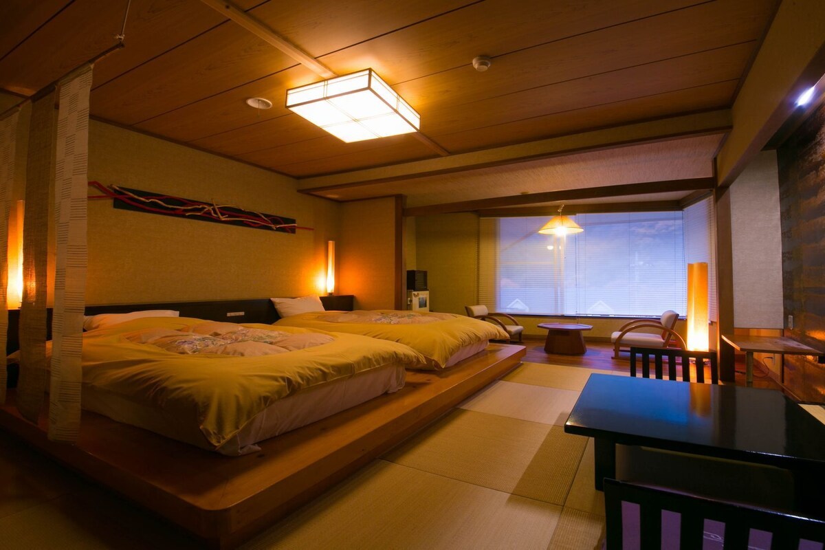 Una habitación tradicional japonesa con suelo de tatami, una cama baja, una pequeña mesa y vistas al exterior a través de un gran ventanal.