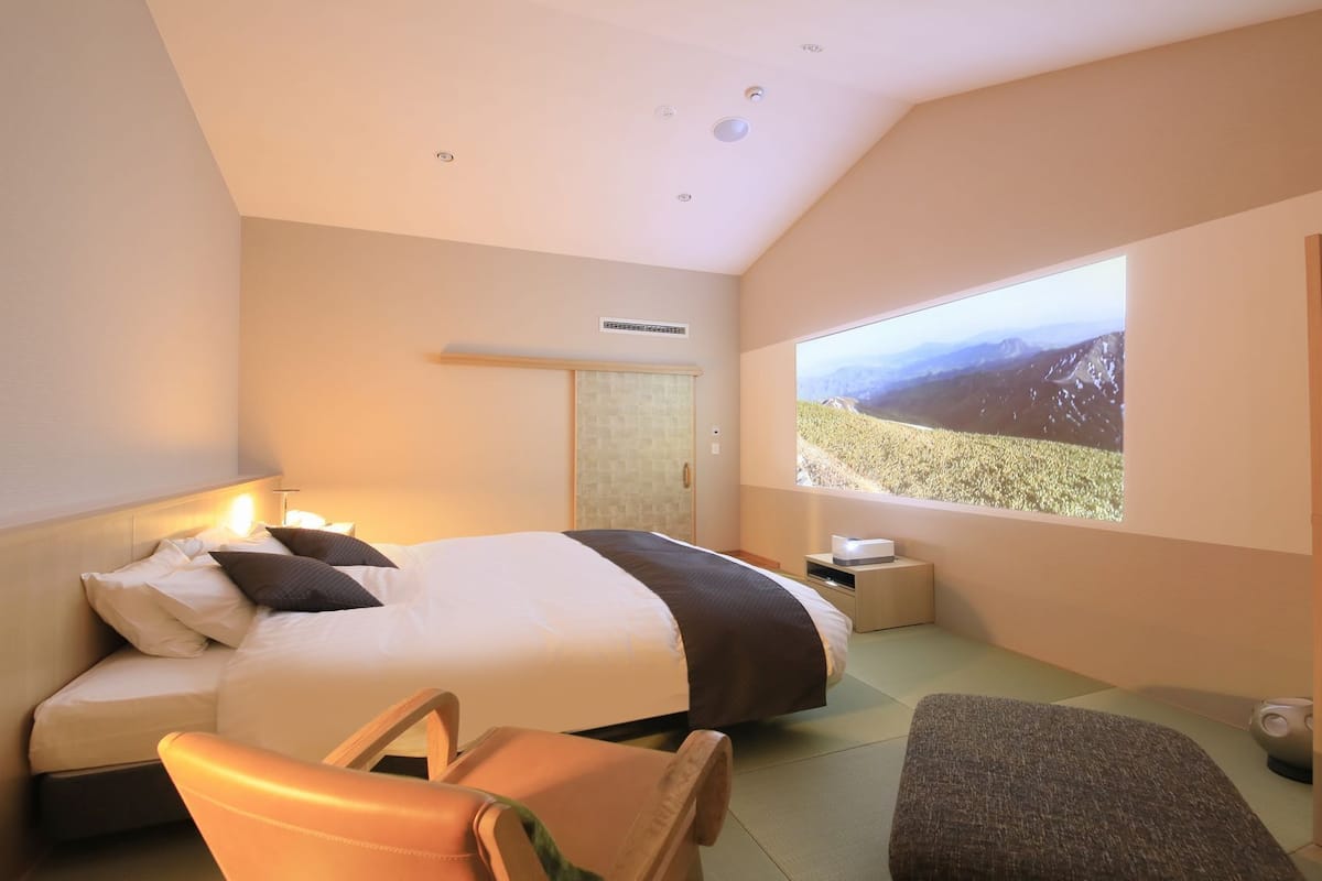 Habitación de hotel moderna con cama, mesita de noche, silla y un amplio ventanal con vistas a las montañas.