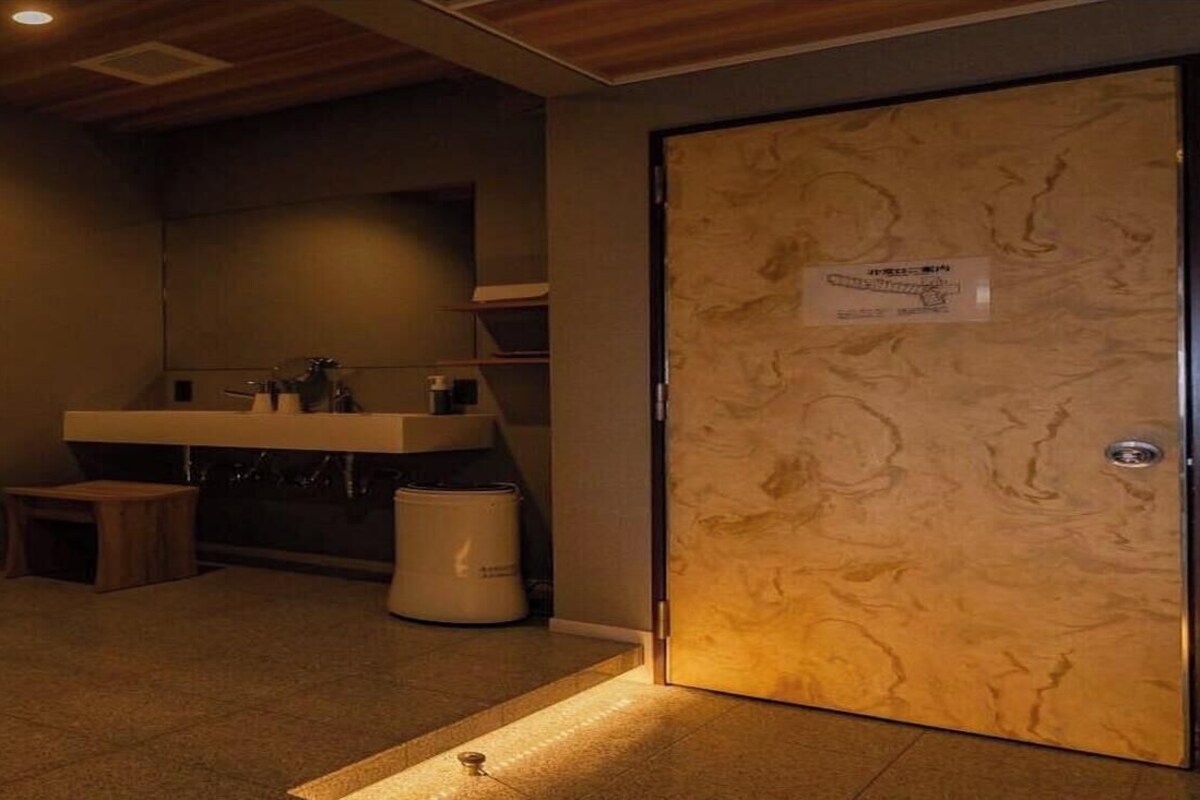 Una habitación con un escritorio de madera, un teléfono, un taburete pequeño, un lavabo y una puerta con un cartel.