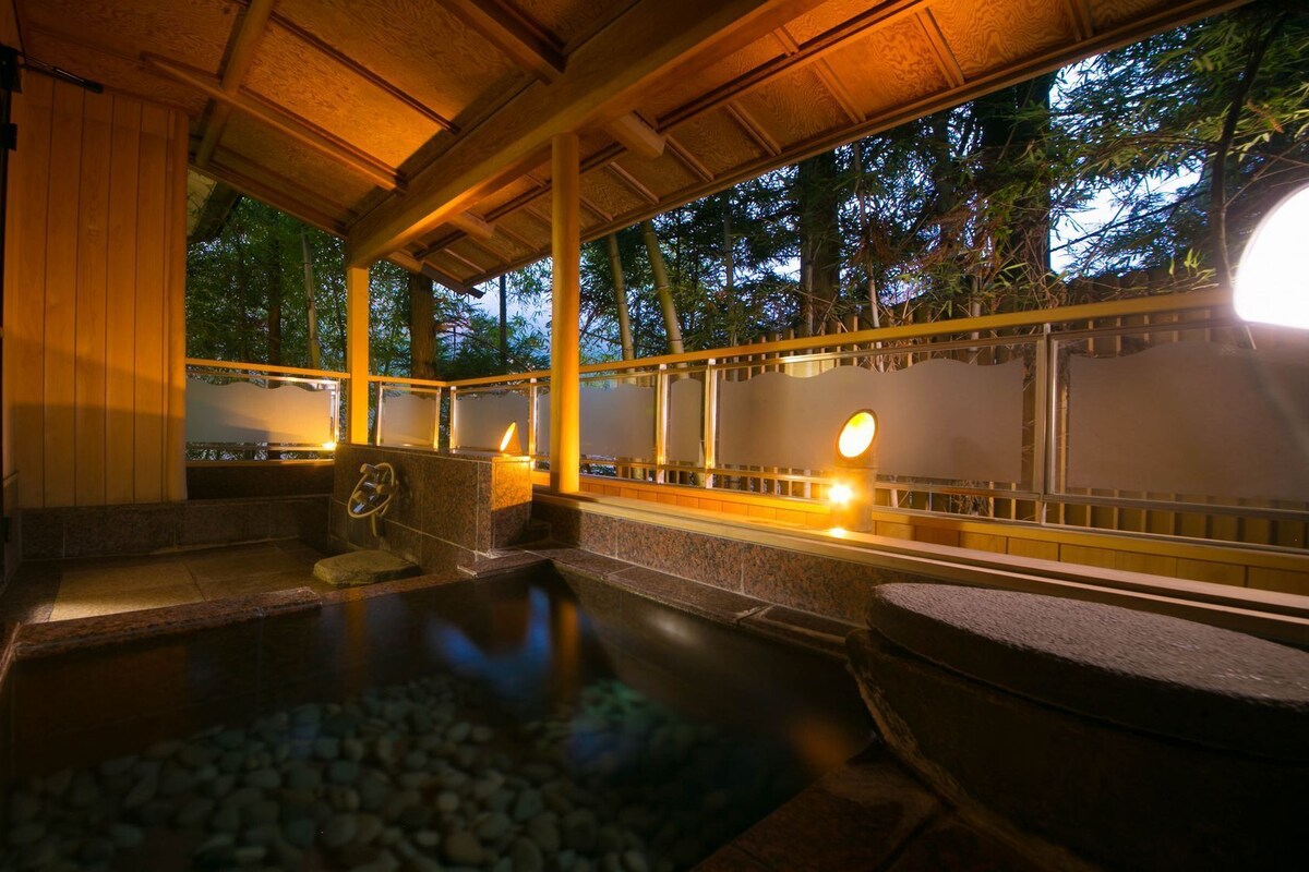 Un onsen japonés tradicional con interior de madera, asientos de piedra y vistas a árboles a través de amplias ventanas.
