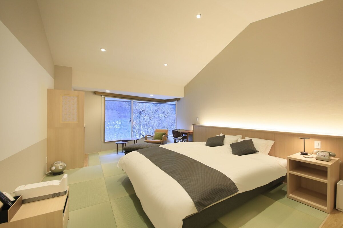 Habitación de hotel moderna con una cama grande, un escritorio, una silla, una mesita de noche con lámpara y vistas a árboles por la ventana.