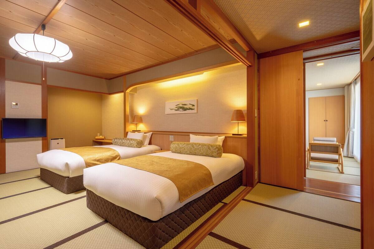 A traditional Japanese-style hotel room with tatami flooring, two beds with matching headboards, a television, and a sliding door leading to a bathroom.