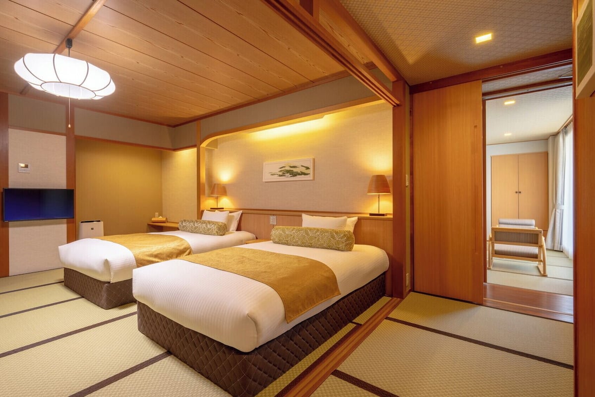 A traditional Japanese-style hotel room with tatami flooring, two beds with matching headboards, a television, and a sliding door leading to a bathroom.