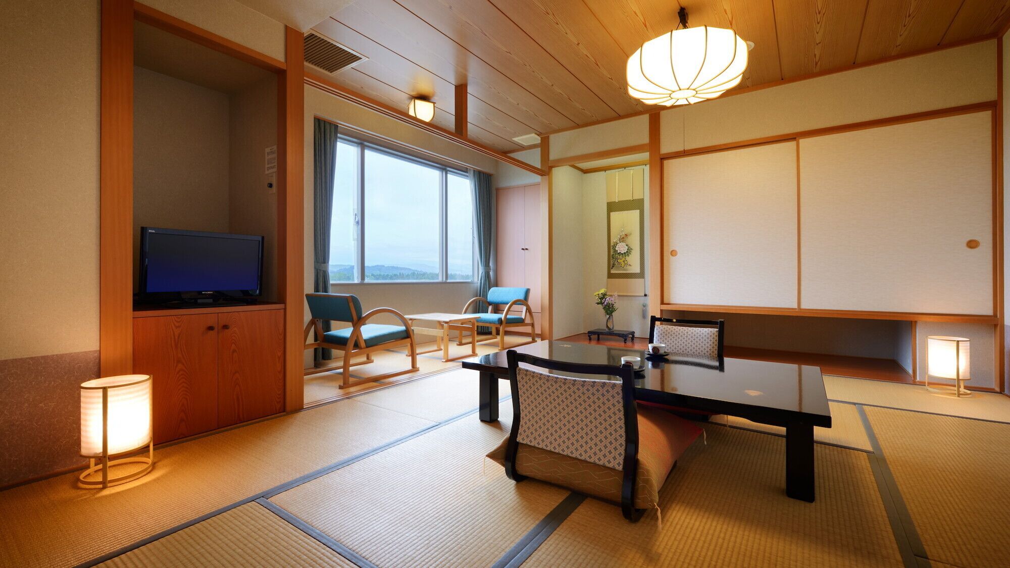 A traditional Japanese-style room with tatami flooring, a television, a dining table with chairs, and a large window offering a view.