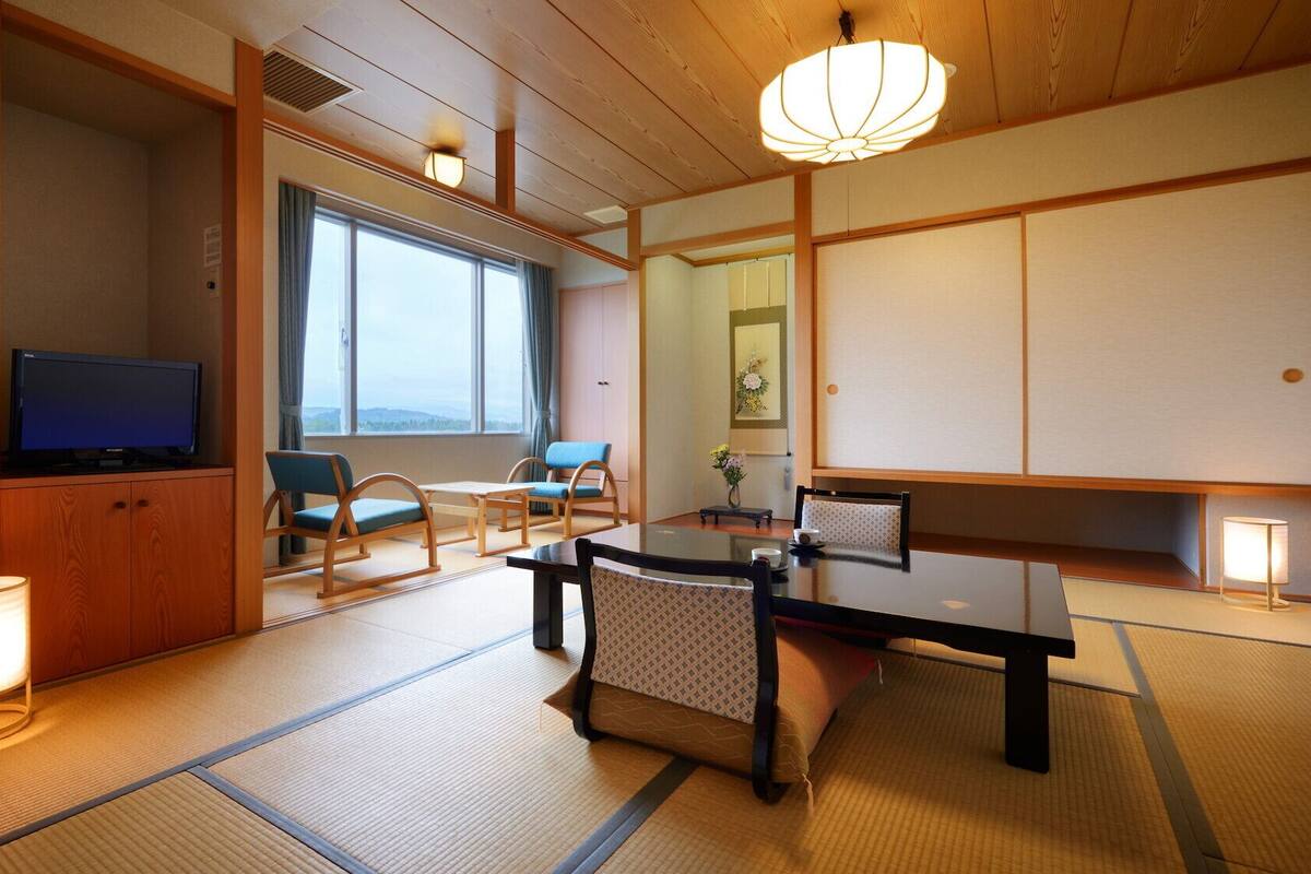 A traditional Japanese-style room with tatami flooring, a television, a dining table with chairs, and a large window offering a view.