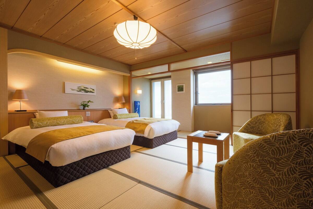 A traditional Japanese-style hotel room with tatami flooring, two beds, a futon, a shoji screen, and a low wooden ceiling.
