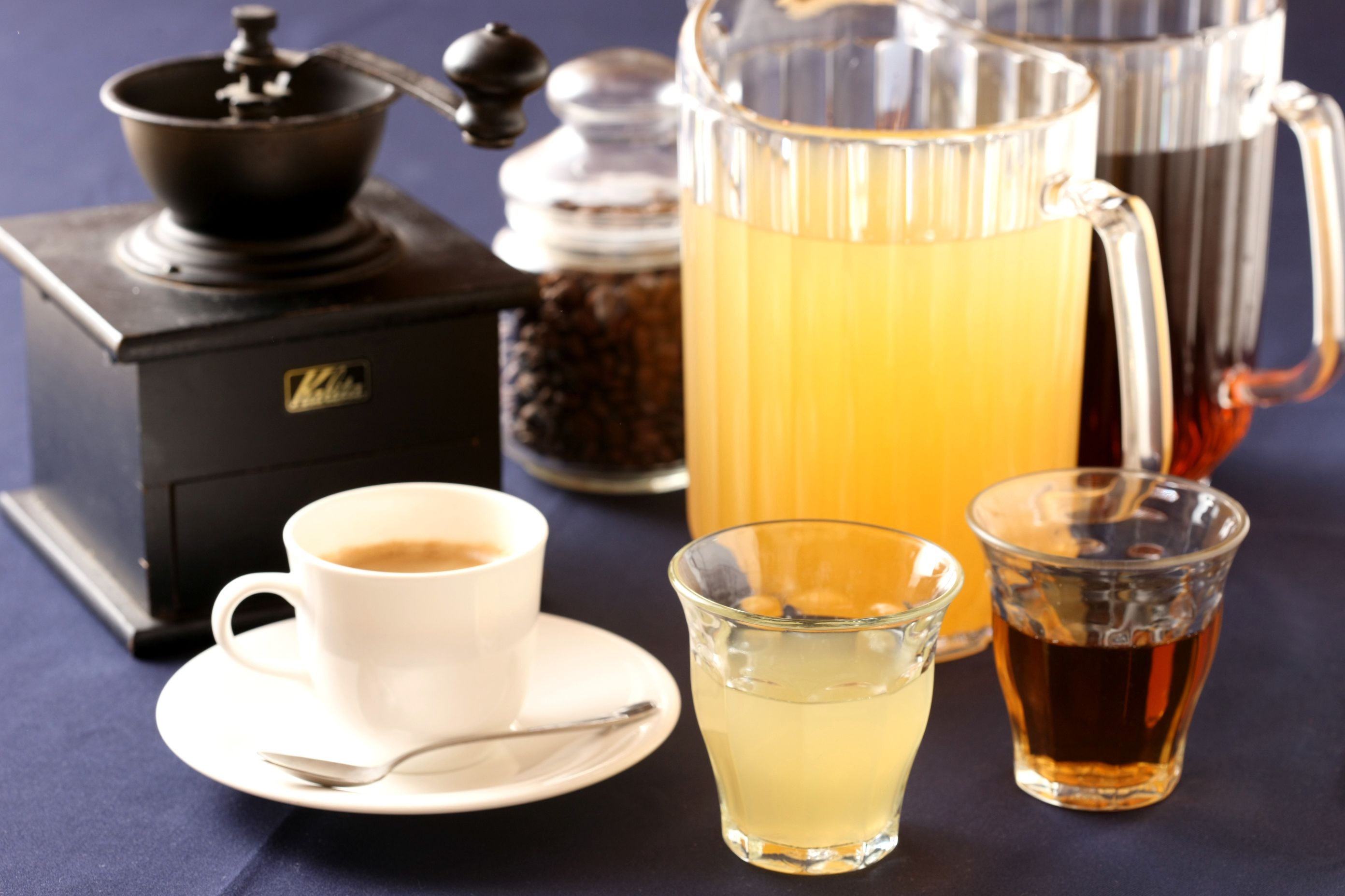 A coffee maker, a coffee cup, a glass of tea, and a glass of lemonade on a table.