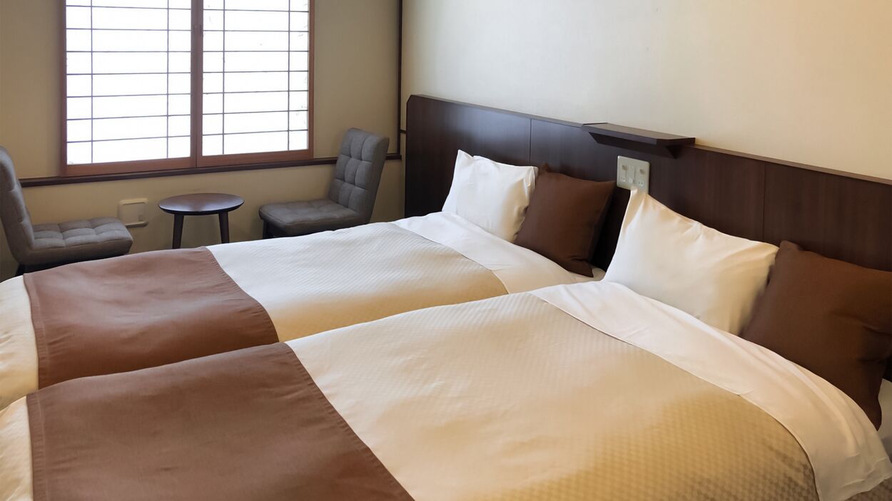 A hotel room with two beds, a small table, a chair, and a window with blinds.