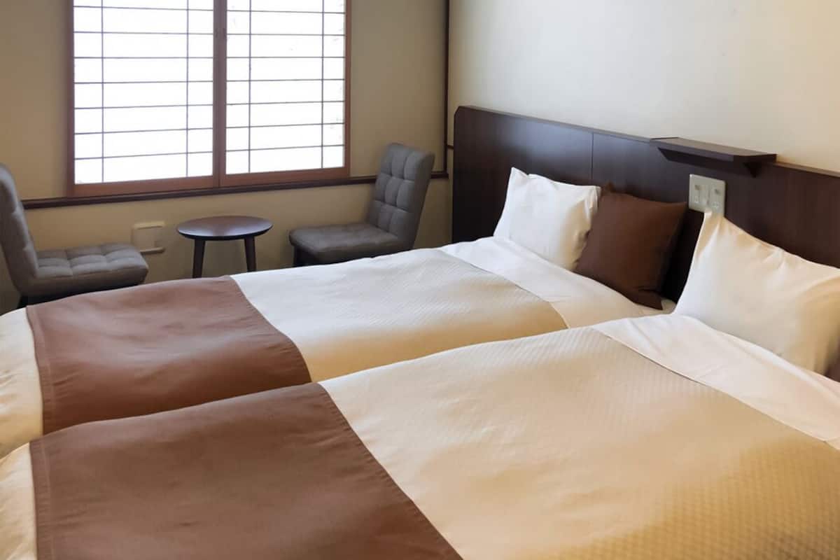 Habitación de hotel con dos camas, una mesita, una silla y una ventana con persianas.