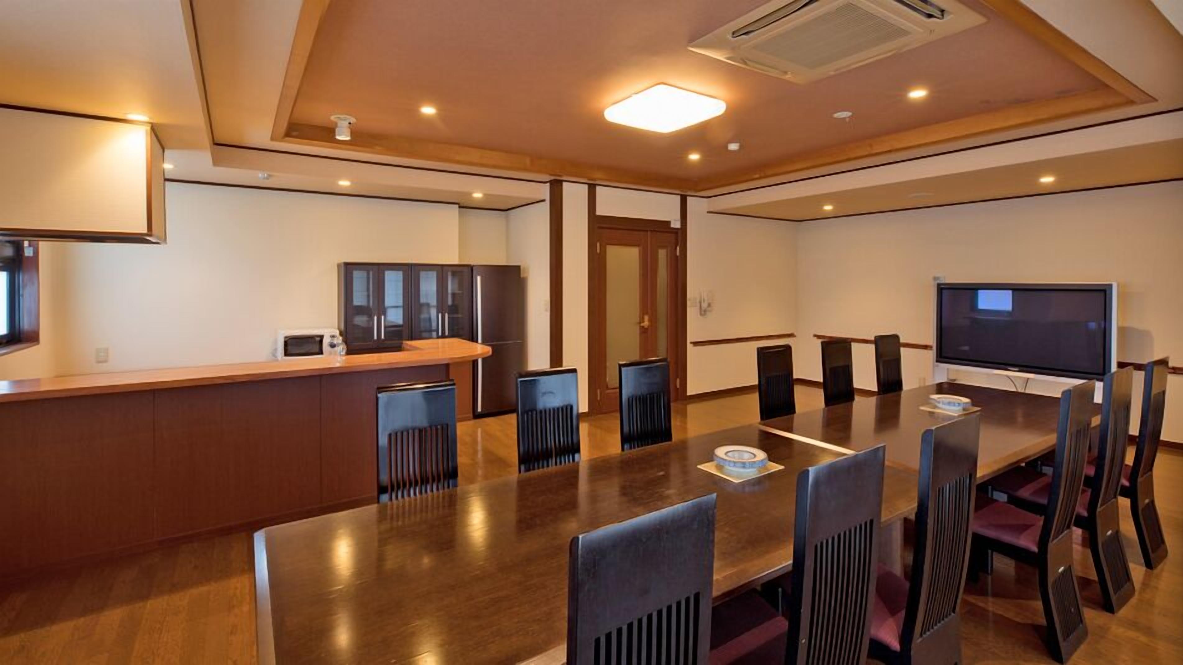 Una sala de conferencias con una larga mesa, sillas, una televisión y una zona de kitchenette.
