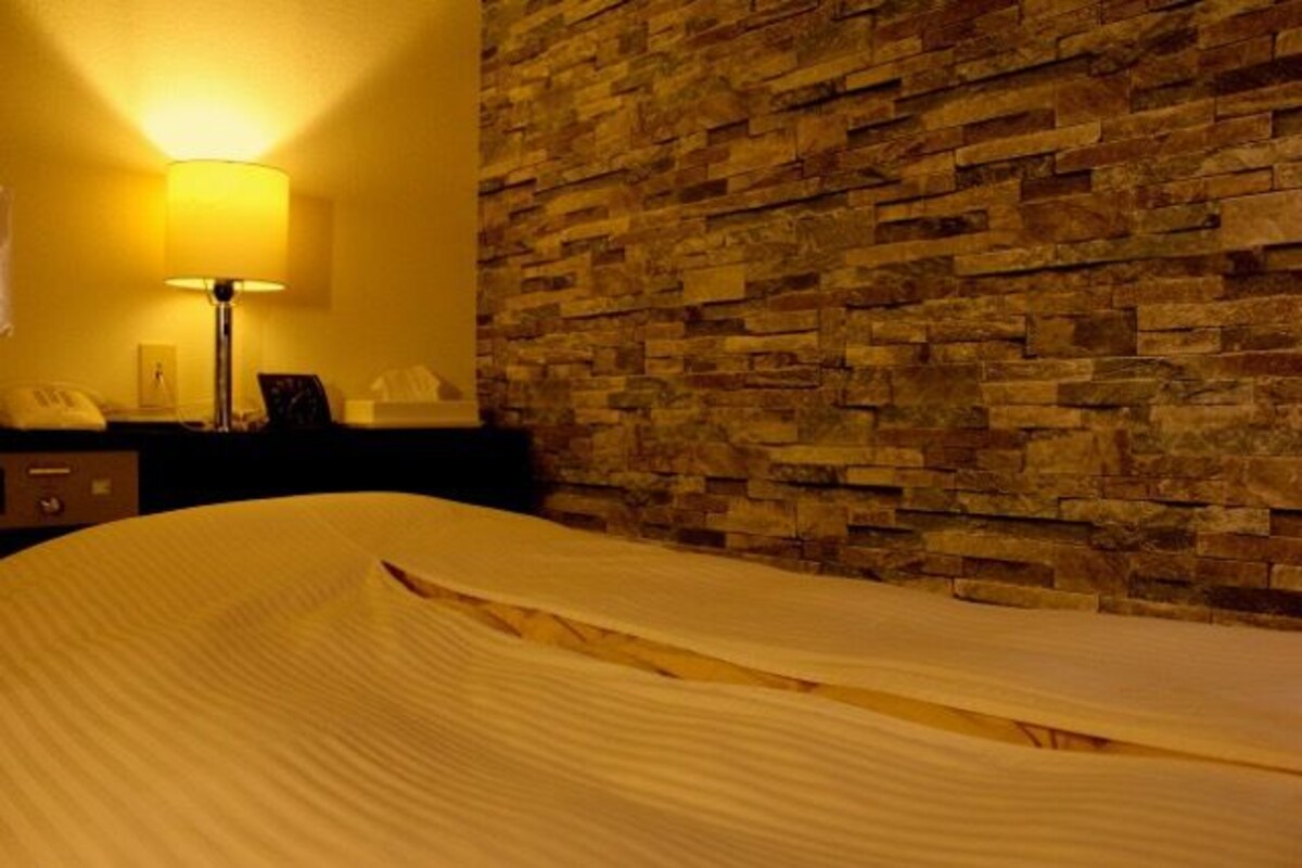 A hotel room with a stone wall, a bedside lamp, a telephone, and a neatly made bed.